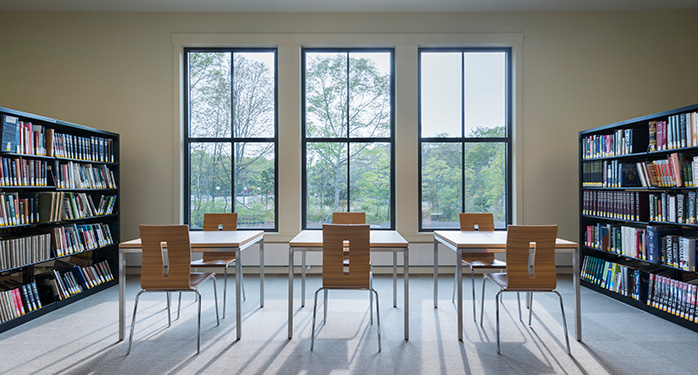 a photo of the back of the library that has two book cases on either side and 3 windows with 3 tables and 6 chairs between them