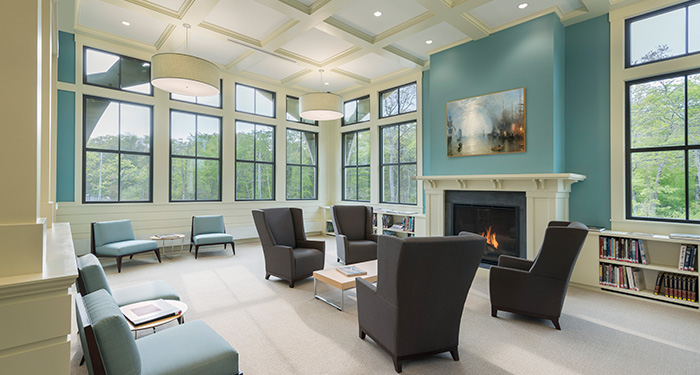 A seating area of the library that has many large chairs in front of a fireplace with a painting hanging over it