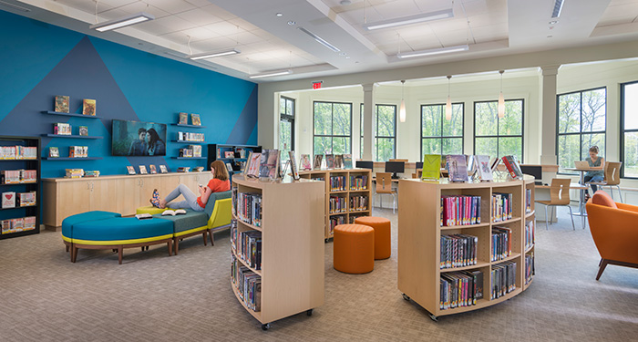 photo of the inside of the teen room of the library picturing the book shelves. seating areas, computers, TV, and two teen girls